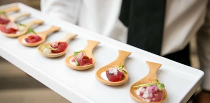 A server holds a white tray lined with spoon-shaped appetizers topped with meat, cream, pickled onions, and microgreens.